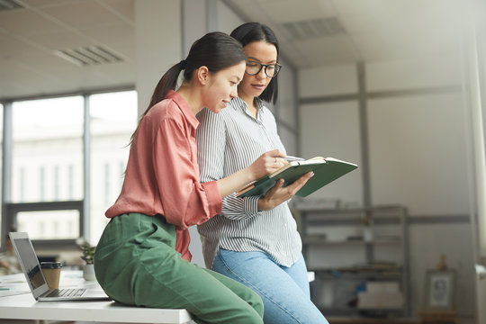 Two young businesswomen in casual clothing holding note pad and looking at their plans for work week together at office - Powered by Adobe