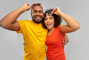 love, relationships and valentines day concept - happy smiling african american couple in heart shaped sunglasses hugging over grey background