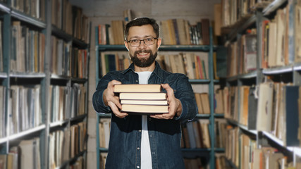Happy bearded man with glasses suggests reading several books, looking at the camera, smiling and holding books forward while standing between bookshelves in the library