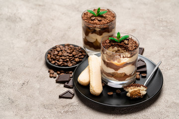 Classic tiramisu dessert in a glass cup, pieces of chocolate and savoiardi cookies on concrete background