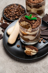 Classic tiramisu dessert in a glass cup, pieces of chocolate and savoiardi cookies on concrete background