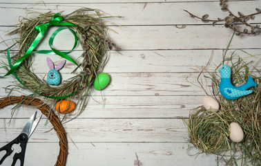 Easter composition. Making an Easter wreath. Willow branches, dry grass, nest, colorful eggs, cute textile bird and Easter Bunny handmade, scissors, ribbons and a bow on a wooden background. 