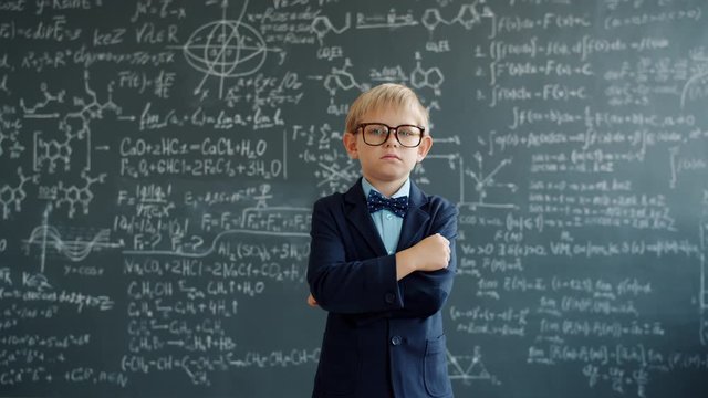 Serious preschooler in suit and glasses is standing with arms crossed in class near blackboard with formulas looking at camera. Education and confidence concept.