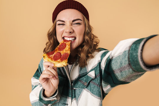 Photo of pretty happy woman in knit hat taking selfie and eating pizza