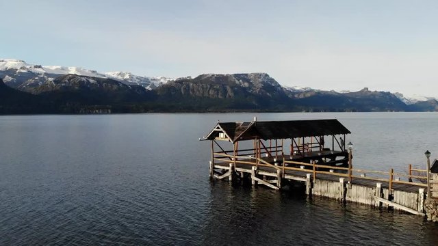 Low fly-over shot of a boathouse on the end of a pier in a calm lake. Majestic snow covered mountains paint the most beautiful background on  a overcast day.