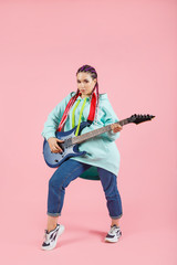 Young guitarist woman in oversize clothes with electric guitar isolated on pink background.