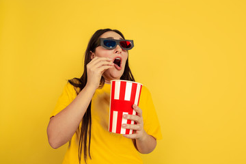 Watching cinema in 3D-eyewear with popcorn. Caucasian woman on yellow studio background. Beautiful brunette model in casual style. Concept of human emotions, facial expression, sales, ad, copyspace.