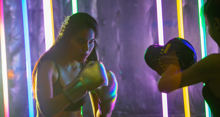 Beautiful 20s Asian Woman trains punching with coach on Silver gold Mitts Gloves. Office Girl exercise at Modern multi color Fashion Neon Muay Thai Boxing Gym with sweat water splash