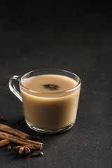 Popular Asian warming drink masala tea in a clear glass cup with coriander, cinnamon and rose stem on a black background.