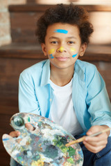 Portrait of African young boy holding palette with paints and looking at camera he painting at studio © AnnaStills