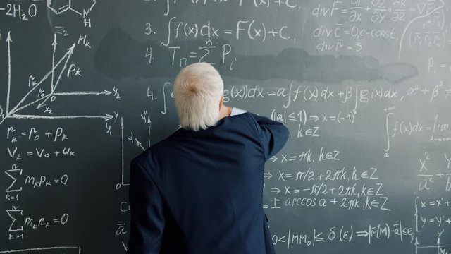 Slow motion of intelligent man in suit writing formulas on blackboard indoors in college class concentrated on science. People and intelligence concept.