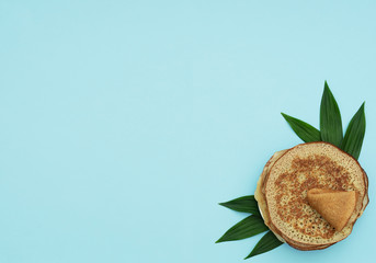 Fototapeta na wymiar Homemade pancakes on blue background with green leaves. Maslenitsa, spring festival concept. Russian traditional food. Pancake week. Delicious breakfast. Flat lay style with copy space.