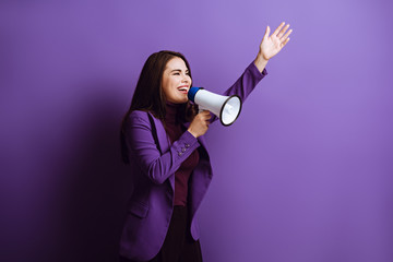 excited young woman talking in megaphone while standing with raised hand on purple background