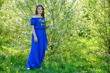 A young woman in a blue dress stands at full length near flowering trees