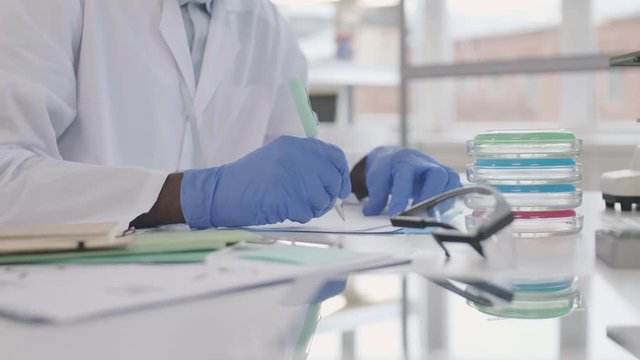 Close up of unrecognizable African doctor sitting at desk in lab and writing in notebook