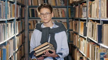 Portrait of a handsome teenage boy in glasses with books in his hands in the library