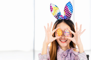 Attractive Asian young woman wearing bunny ears holding up colorful easter eggs in front of her eyes with a toothy smiling happy face. Holidays, tradition and people concept with copy space.