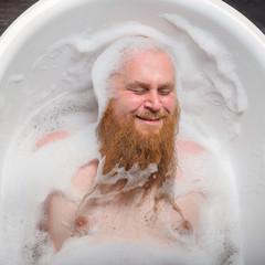 Portrait of a bald man with a long red beard takes a bath with foam. Top view on a cheerful funny...