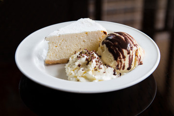 A slice of cheese cake, ice cream, and whipped cream on a white plate and black background.
