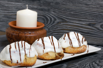 Cinnabons in white glaze, decorated with chocolate. Lie on a plate. Nearby is a white candle in a clay candlestick. Against the background of brushed pine boards.