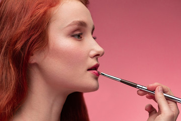 Makeup artist applies lipstick. Hand of make-up master, painting lips of young redhead girl....