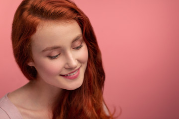 Fototapeta premium Portrait of smiling redhead girl. Perfect skin. Beautiful female model with long hair over pink background. Young cute woman with clear skin. Skincare and facial treatment concept.
