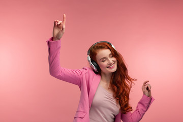 Enjoy listening to music. Beautiful young redhead woman in headphones listening music. Funny smiling girl in earphones and pink jacket dancing on pink background.