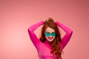 Portrait of smiling redhead girl. Perfect skin. Beautiful female model with long hair over pink background. Young cute woman in green sunglasses with clear skin touching her hair. Copy space.