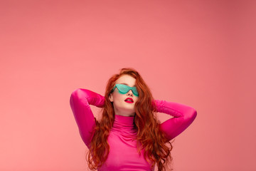 Portrait of smiling redhead girl. Perfect skin. Beautiful female model with long hair over pink background. Young cute woman in green sunglasses with clear skin touching her hair. Copy space.