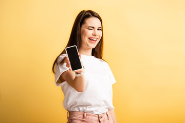 cheerful girl winking at camera while showing smartphone with blank screen isolated on yellow