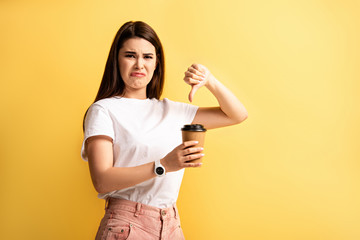 displeased girl showing thumb down and grimacing while holding coffee to go isolated on yellow