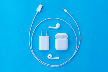 Top view flat lay plastic white wireless headphones for smartphone, charging case, Lighning USB, adapter on a pastel blue background. Copy space.