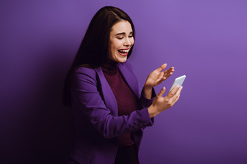 excited girl standing with open arm and laughing while having video chat on purple background