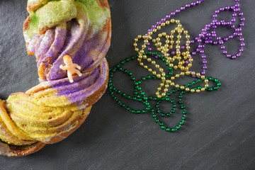Flay lay of a festive Mardi Gras King Cake with purple, gold and green beads with a small plastic baby on the King Cake; tradition; gray slate background