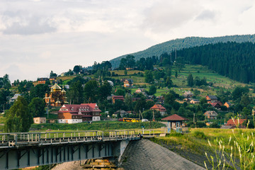Fototapeta na wymiar Ukrainian village in the Carpathians in sunny evening. Beautiful landscape during the evening light. Summer country side view. Authentic abandoned railway with traditional houses, church, hills.