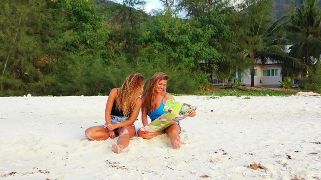 Excited young women reviewing travel guide on unspoiled beach. Summer vacation on exotic islands