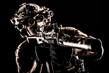 Army special operations soldier, commando fighter in full tactical ammunition, helmet with radio headset and night vision device, aiming short barrel assault rifle in darkness, low key studio shoot