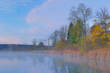 Fototapeta premium Foggy, autumn landscape of the shoreline of Whitford Lake at dawn with reflections in calm water, Fort Custer State Park, Michigan, USA