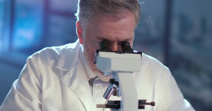 A mature bearded Caucasian male researcher or technician wearing protective gloves and working with a microscope makes some notes about his findings.