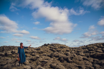 Young beautiful woman in blue dress stands on the volcanic formations covered by green moss on a background sky. Place for text or advertising