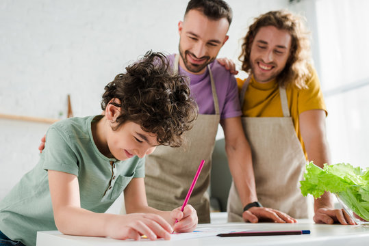 selective focus of mixed race kid drawing picture near happy homosexual parents