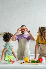 bearded homosexual man covering eyes with cherry tomatoes near mixed race son and partner