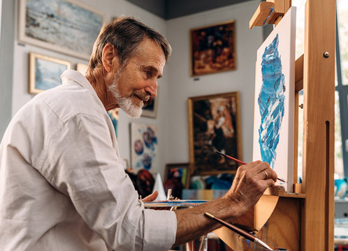 Bearded artist sitting in front of easel and signing abstract picture with a paintbrush