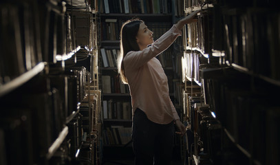 Concentrated girl looking for book in bookcase in dark library standing between rows of shelves © MYDAYcontent