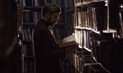 Man consumed with reading in dark library standing among rows of shelves with books, side view