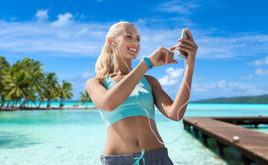 fitness, sport and people concept - smiling young woman with smartphone and earphones listening to music and exercising over wooden pier, palm trees and sea in french polynesia on background