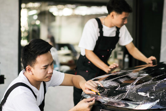Two Asian detailing film paint protection men specialist cutting and wrapping car in garage showroom, protective coating installation and polishing automobile for vehicle care service business concept
