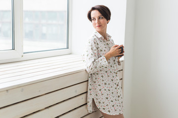 Calm and coziness. Beautiful young woman with cup of tea standing near the window at home