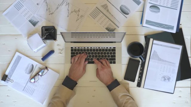 Male arms of businessman typing text on laptop keyboard at white desk. Top view hands of manager working on notebook in office. Diagrams with statistics at background. Concept of business lifestyle