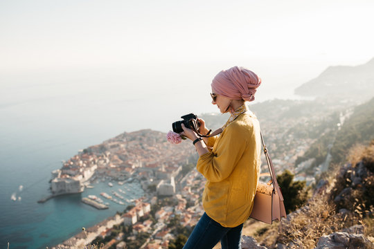 Woman with hijab standing above old city of Dubrovnik and taking photos with her camera. She is happy and relaxed. She is enjoying sunset. 